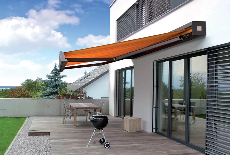 Ausgefahrene Gelenkarmmarkise in Orange an einer weißen Fassade über einer Holzterrasse mit Grill