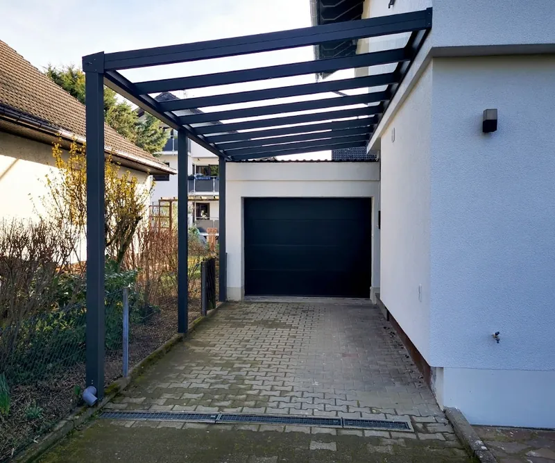 Schmaler Carport mit Pultdach und dunkelblauen Stützen über einer Zufahrt zur Garage