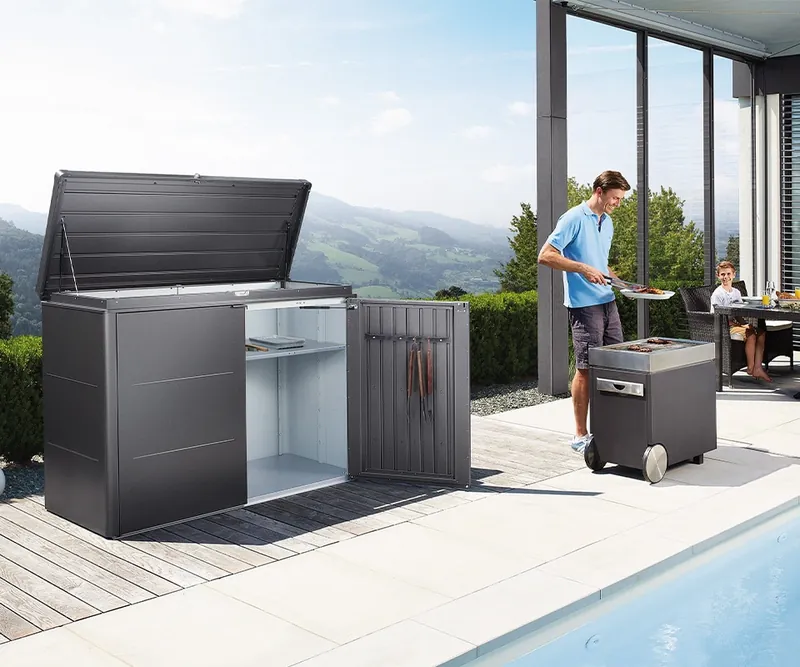 Dunkelgraues Stauraum-Highboard mit geöffnetem Deckel auf einer Holzterrasse am Pool, daneben ein Mann am Grill