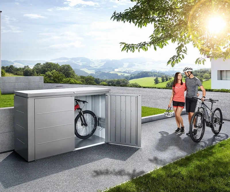 Silberfarbenes Highboard mit geöffneter Tür und darin abgestelltem Fahrrad, daneben ein Radfahrerpaar