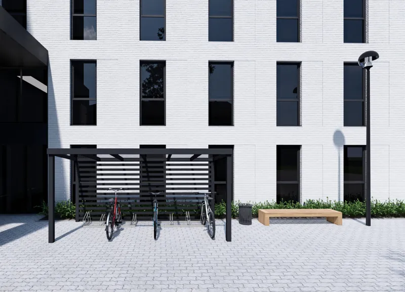Fahrradüberdachung mit schwarzem Rahmen und Lamellenrückwand vor einem weißen Bürogebäude, darunter drei abgestellte Fahrräder, daneben Bank und Laterne