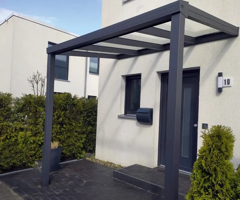 Anthrazitfarbenes Vordach mit opalweißer Eindeckung vor einer Haustür mit Hausnummer und Briefkasten, daneben eine Hecke