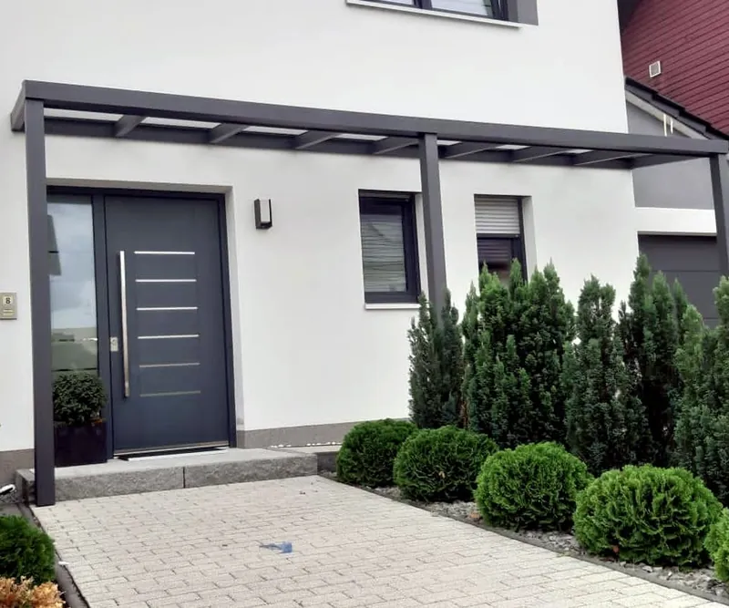 Breites flaches Vordach über einer dunklen Haustür mit Edelstahlgriff, davor Thujen und Pflaster