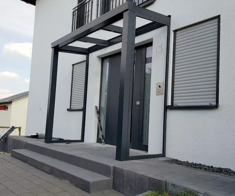 Vordach auf vier anthrazitfarbenen Stützen über einer Haustür mit Natursteinstufen und Rollläden an den Fenstern