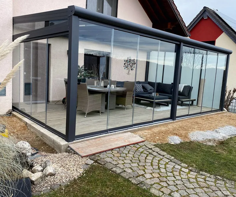 Verglaster Terrassenanbau mit anthrazitfarbenen Rahmen und Glasschiebeelementen, Lounge-Möbel im Innenraum, Rasen und Pflasterweg davor